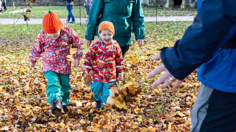 Two young children in water proof coats and pumpkins hats are kicking colourful autumn leaves in the grass and smiling.